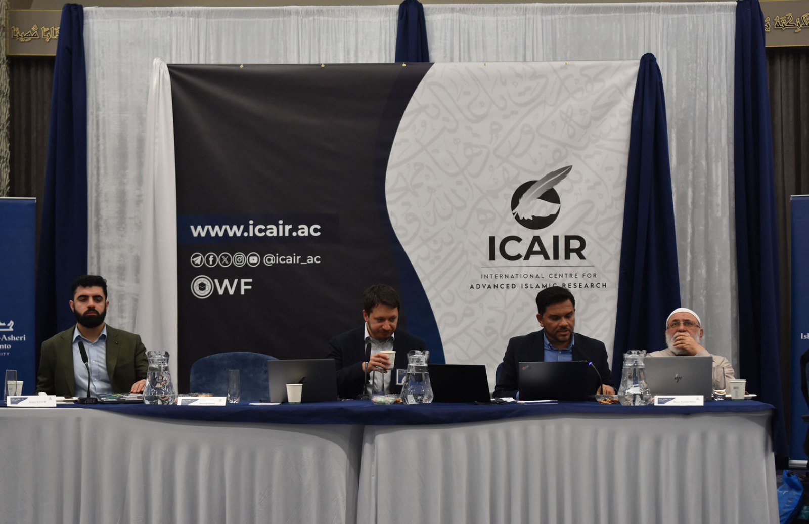 Four panellists seated at the symposium table beneath ICAIR's backdrop at the Jaffari Community Centre.