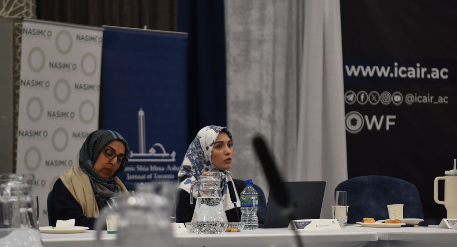 Two participants at the symposium table, one speaking, with NASIMCO and ICAIR banners behind them.