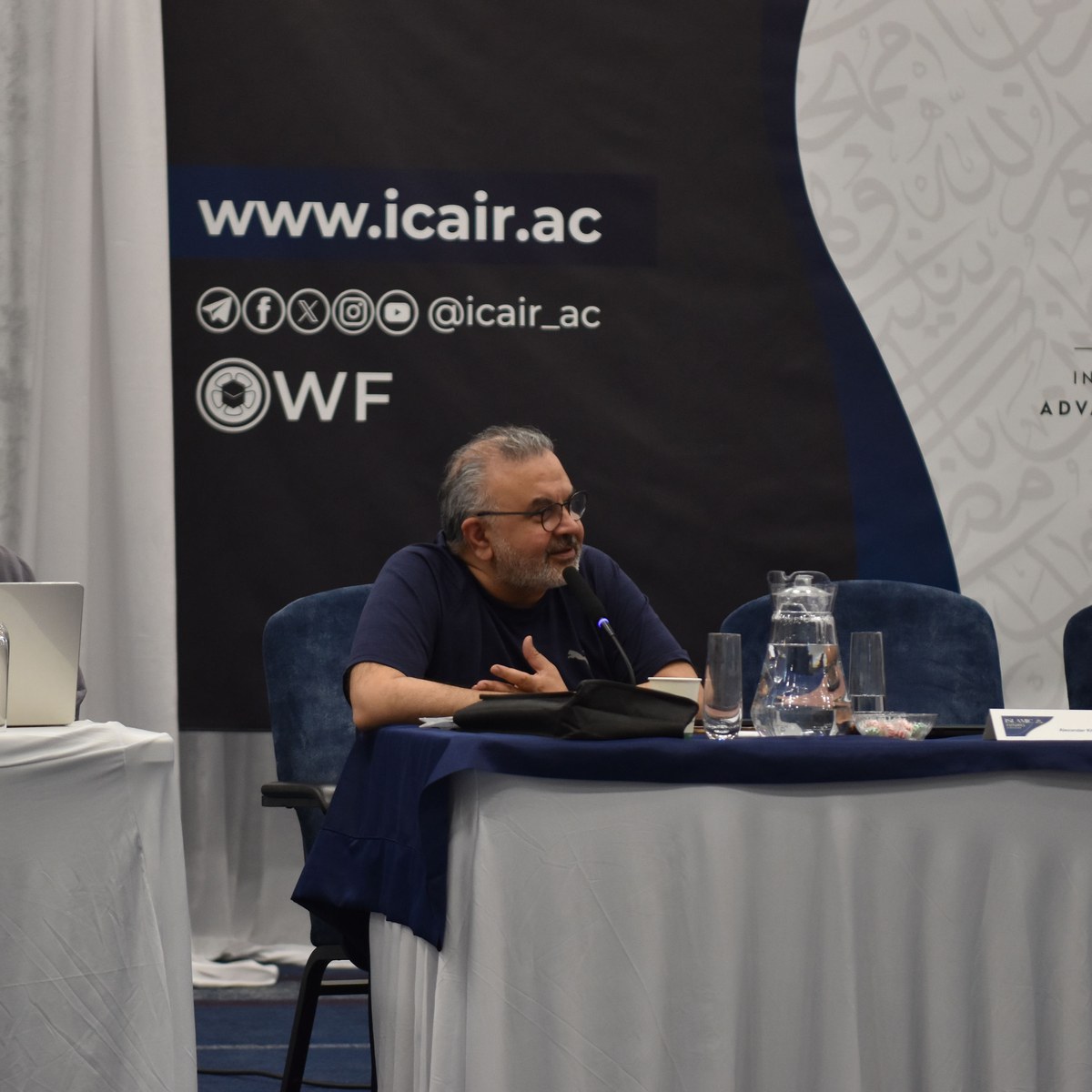 A speaker at the table during an ICAIR session, with the ICAIR backdrop running behind him.