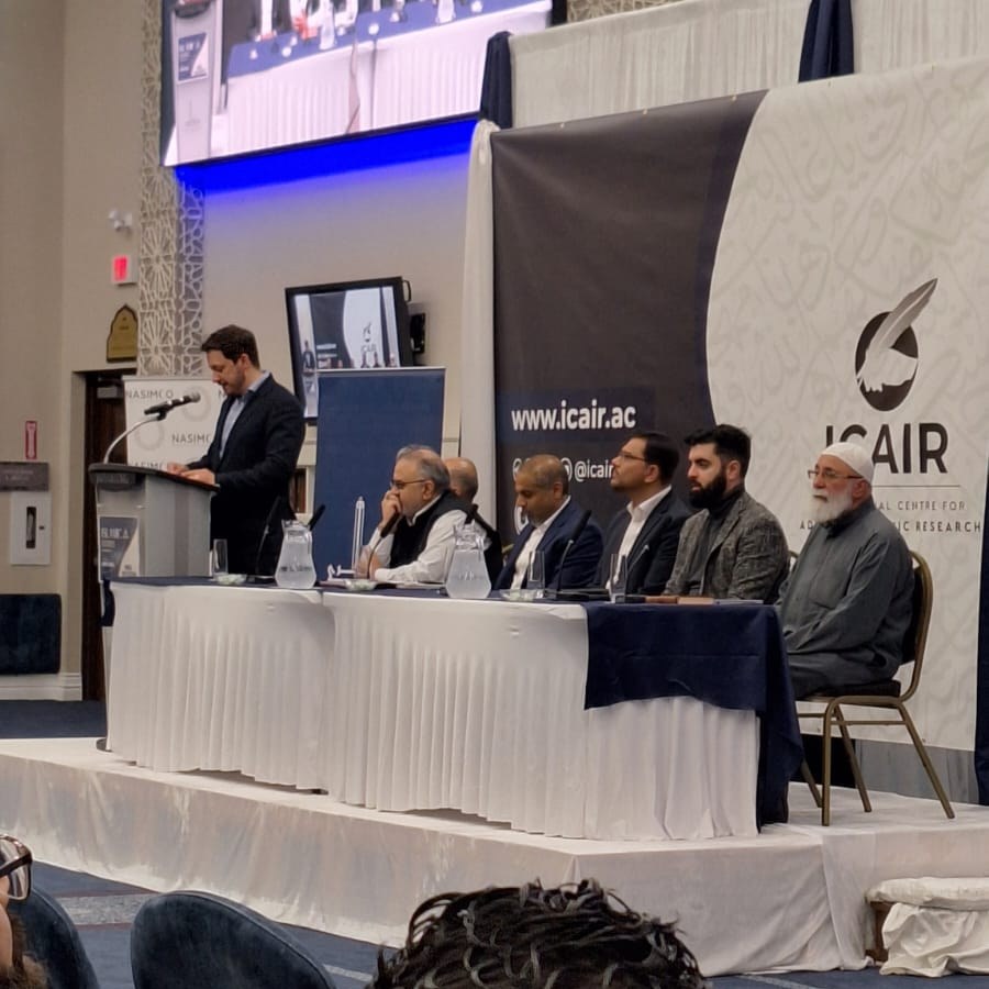 A panel discussion on stage in front of an ICAIR backdrop, with a moderator speaking at the lectern and the panellists seated at a long table.