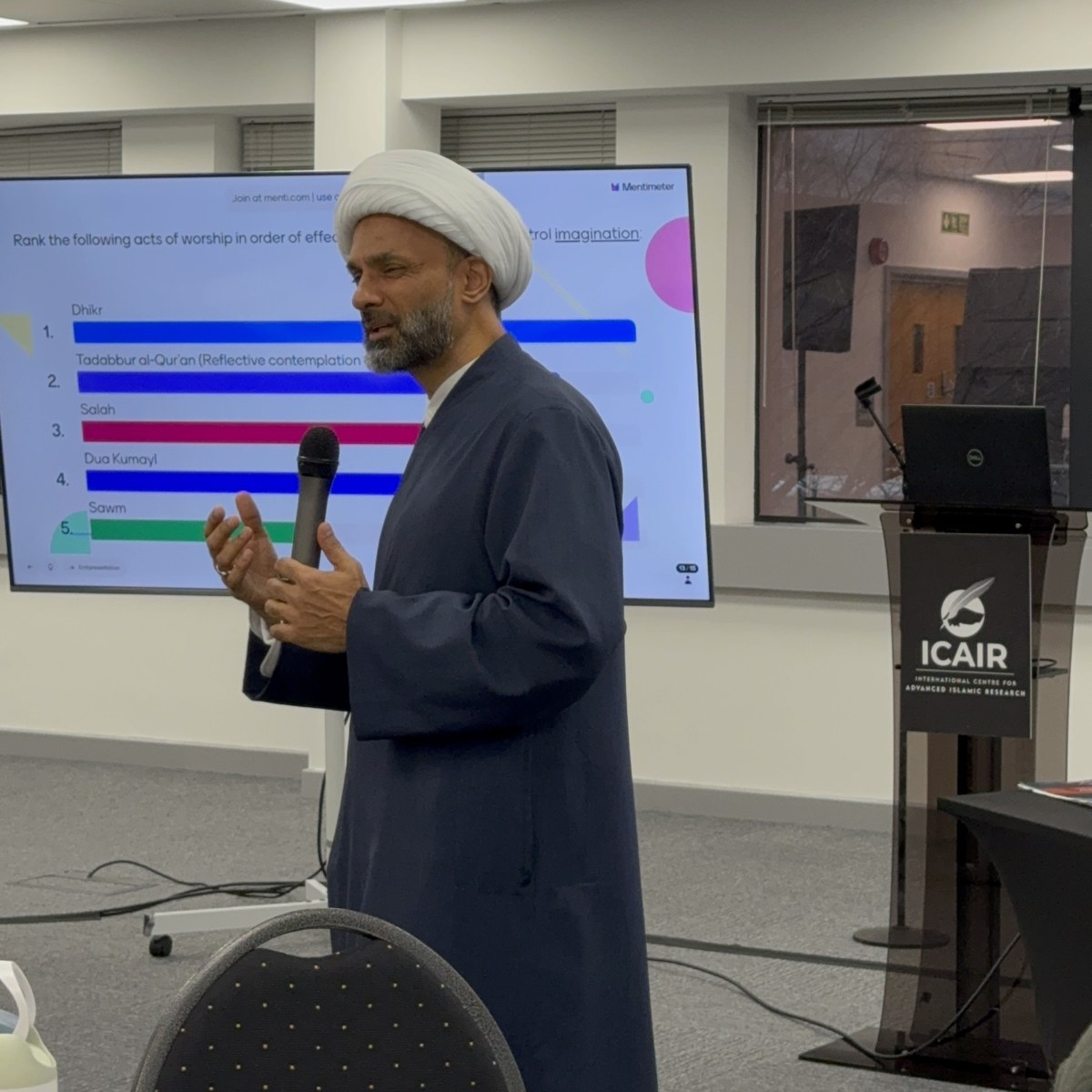 A speaker addressing the workshop with a microphone, with a live audience poll displayed on the screen behind.
