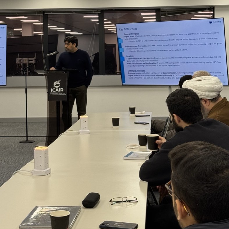 A speaker at the ICAIR lectern during the seminar on cryptocurrency and digital assets, with a slide comparing types of digital asset on the screen beside him.